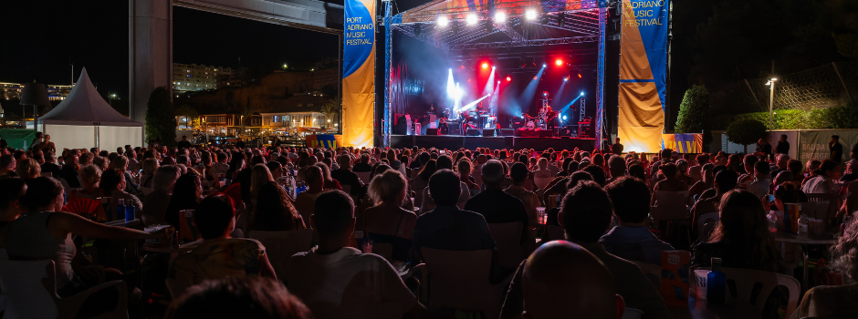 "Large audience seated at Port Adriano watching a live evening concert on an open-air stage with bright red and white lights."
