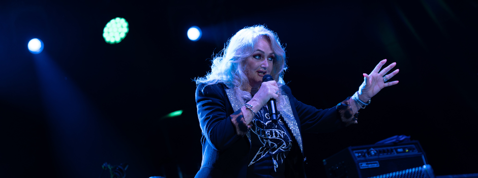 "Bonnie Tyler performing on stage at Port Adriano, holding a microphone and gesturing to the crowd under blue stage lighting."
