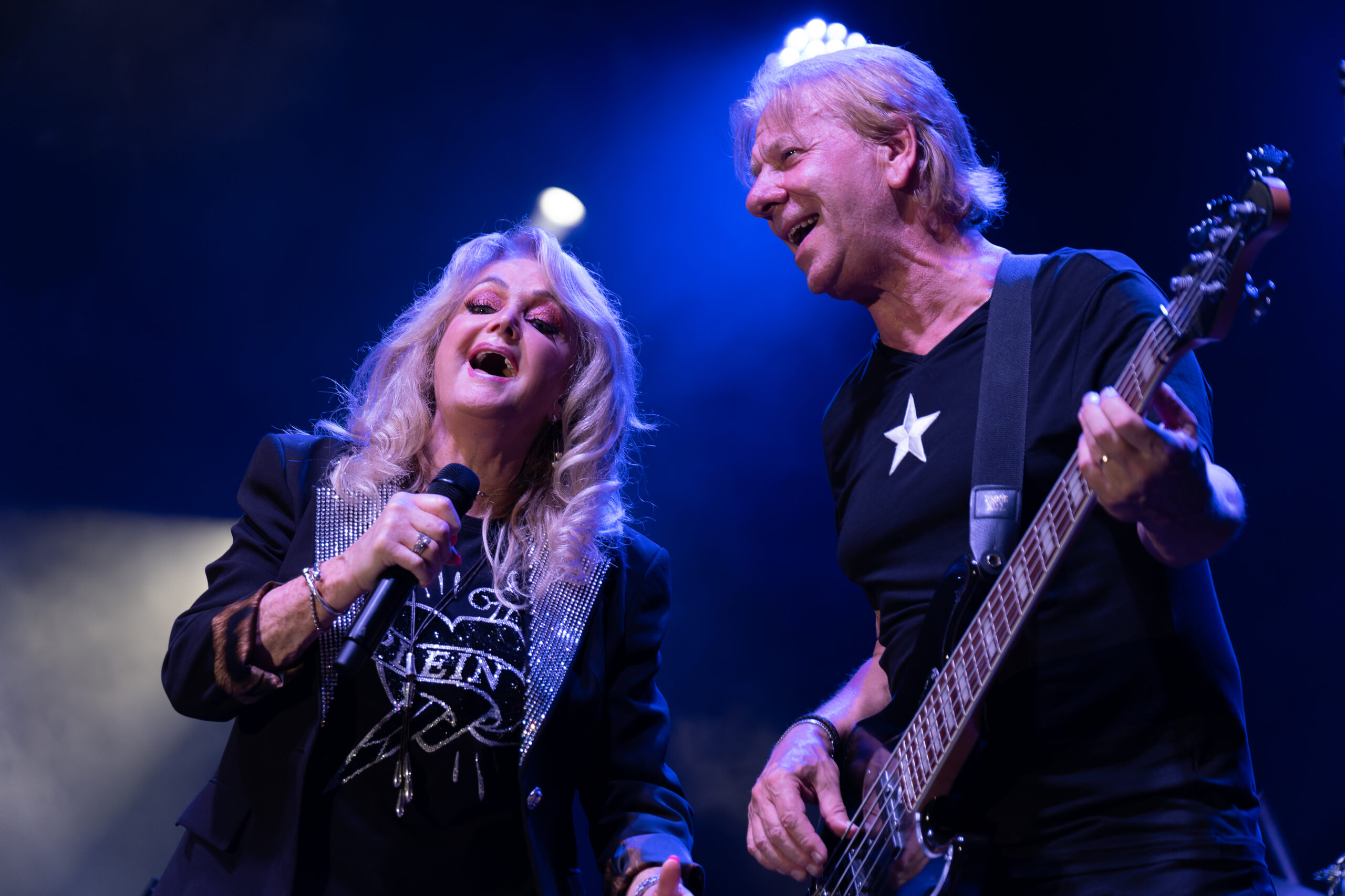 “Bonnie Tyler performing with her bassist at Port Adriano Music Festival, both smiling and singing under blue stage lights.”