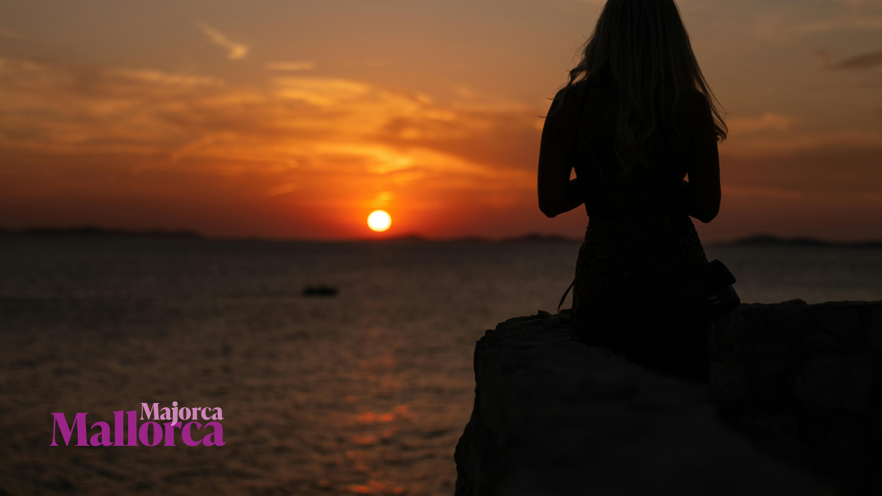 Silhouette of a woman watching the sunset over the sea in Mallorca, with the Majorca Mallorca logo in the lower left corner.